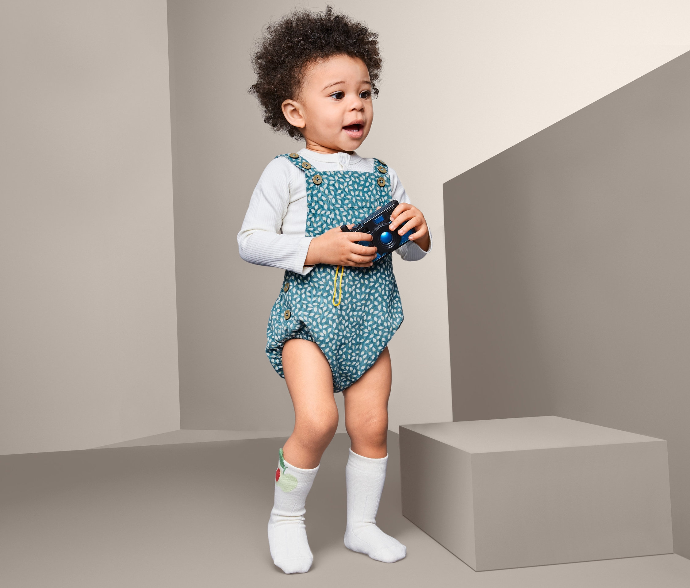 Ein lockiges Kind steht mit einer Spielzeugkamera und trägt einen Feincordstrampler.