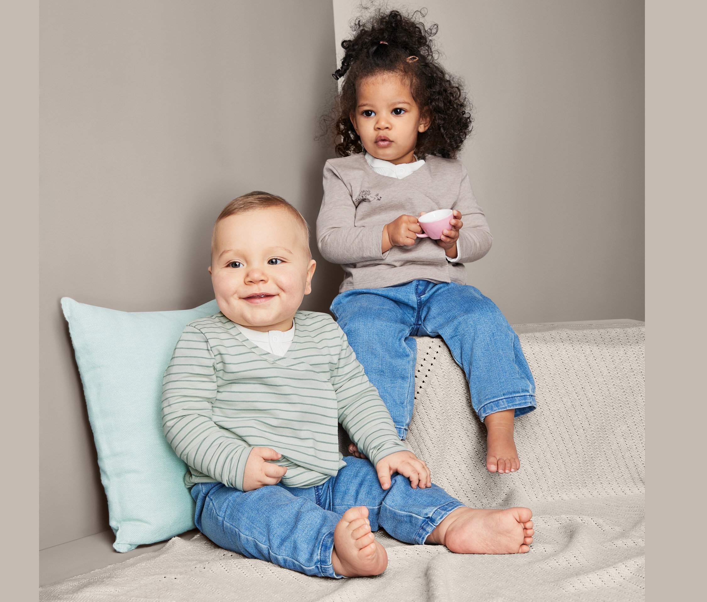 Ein Baby sitzt neben einem Kleinkind. Das Kleinkind hält eine rosa Tasse. Beide tragen Jeans und 2 Baumwoll-Jersey-Wickeljacken.
