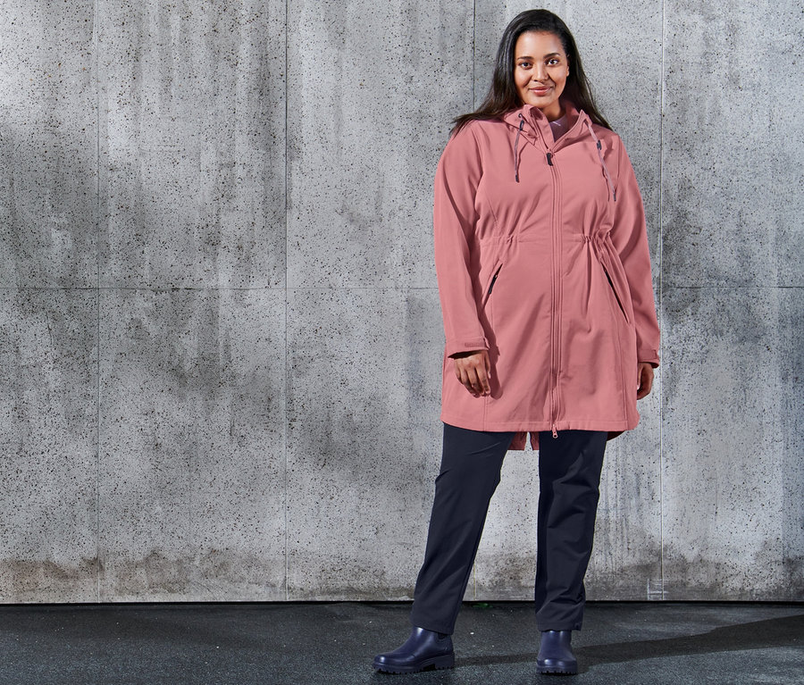 Eine Frau posiert vor einer Betonwand und trägt einen Softshell-Mantel und eine dunkelblaue Softshell-Joggpants.