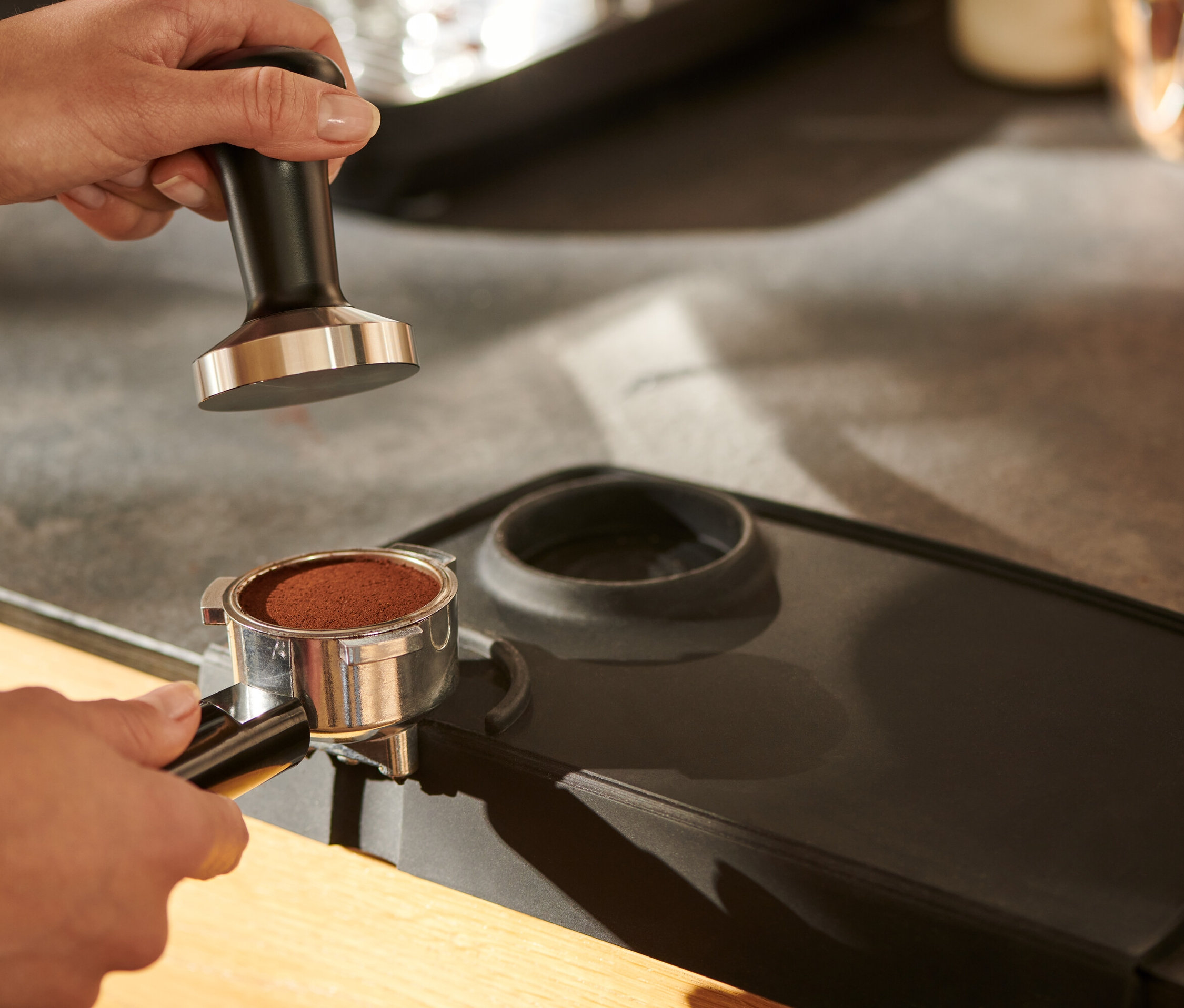 Eine Hand hält einen Tamper, schwarz, über einer Tampermatte.