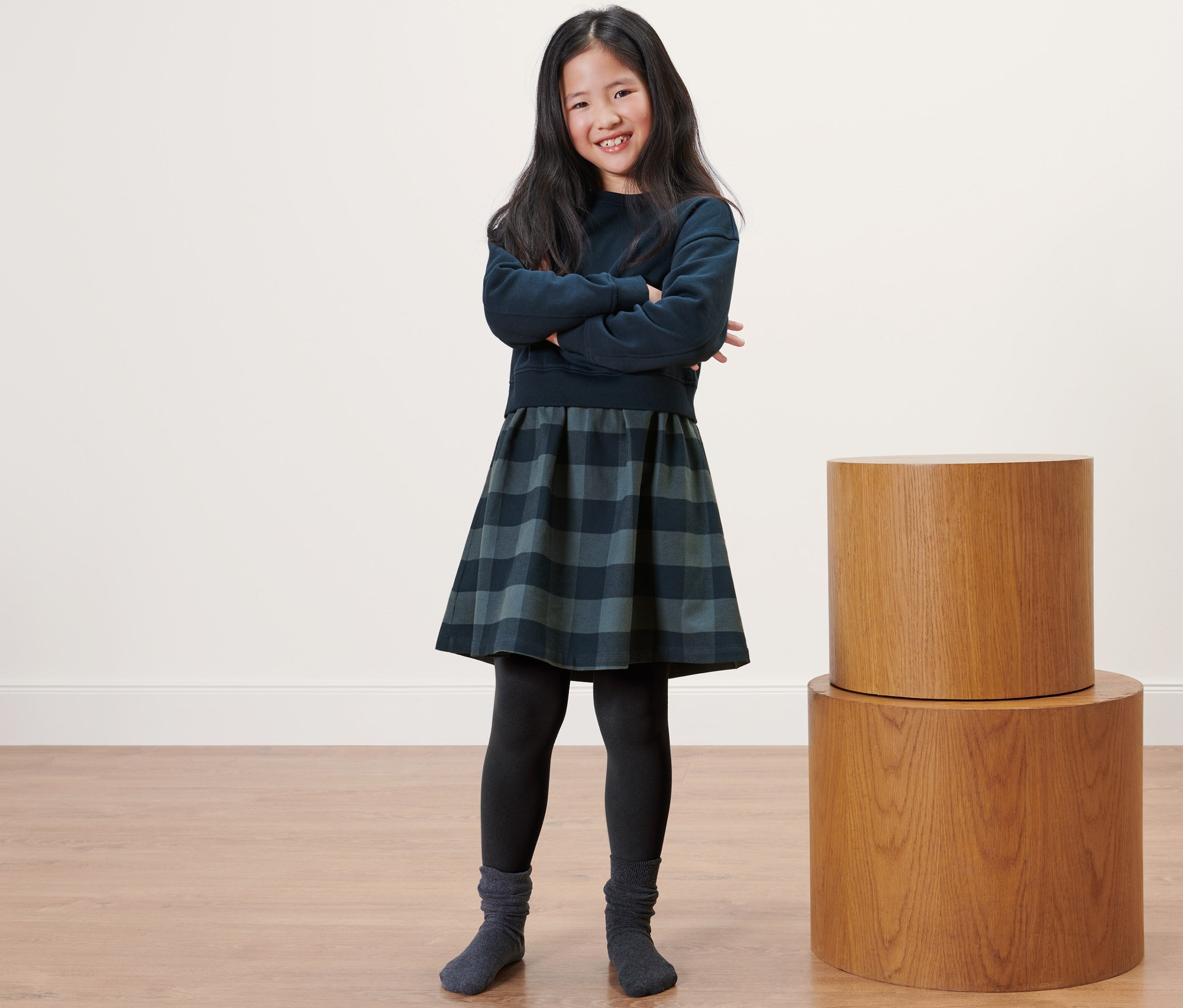 Lächelndes Mädchen posiert mit verschränkten Armen und trägt Kinder-Thermoleggings, einen Pullover und einen karierten Rock.