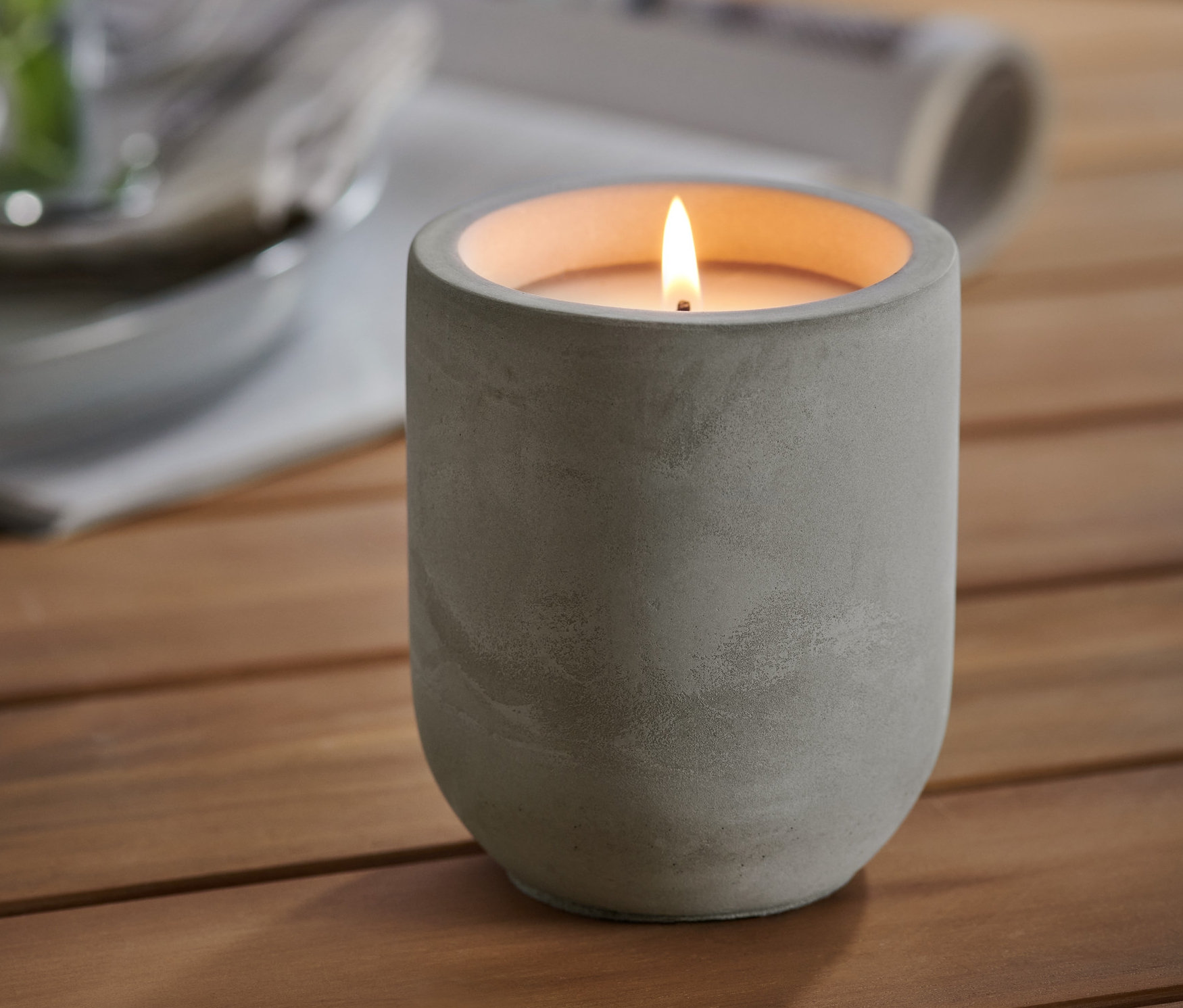 Eine brennende Citronella-Kerze in einem ZementbehÀlter steht auf einem Holztisch. Im Hintergrund befinden sich ein Glas mit einer Pflanze und Teller.
