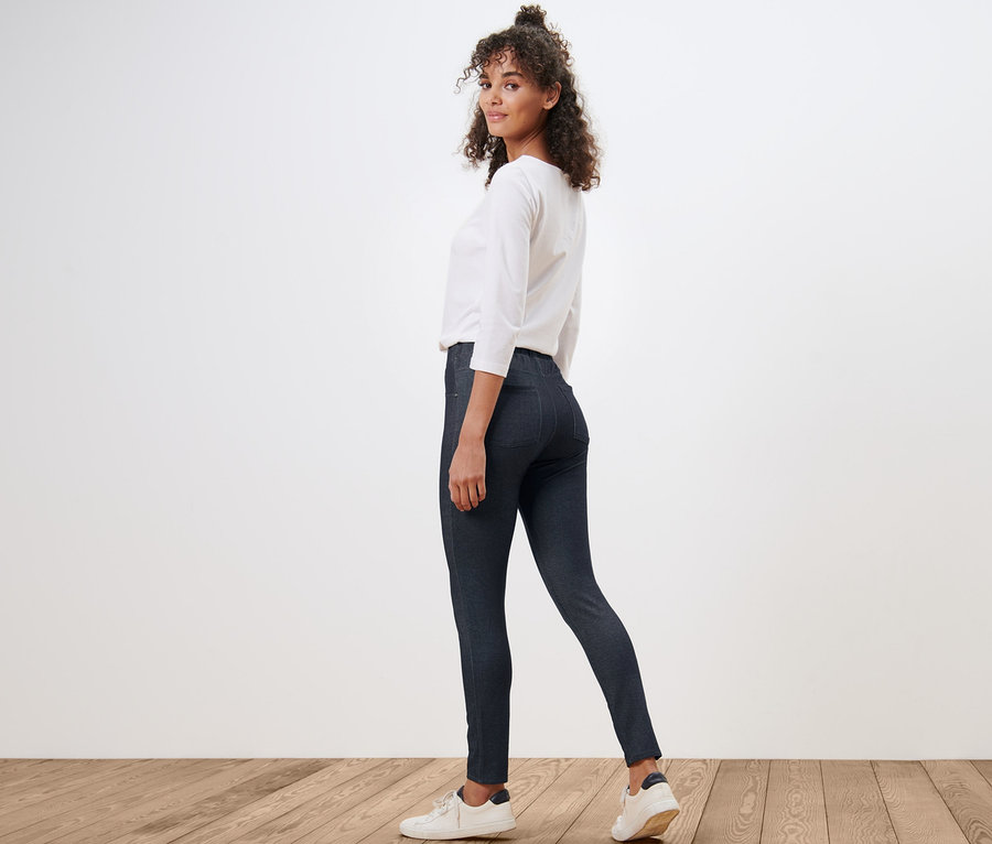 Frau blickt über die Schulter und trägt Leggings in Jeansoptik, eine weiße Bluse und Turnschuhe.