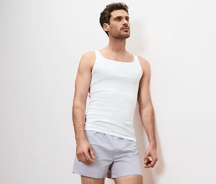 Mann lehnt an einer Wand und trägt ein weißes Unterhemd und graue Shorts. Das Unterhemd ist aus 2 Qualitäts-Feinripp-Unterhemden.