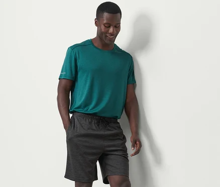 Mann in grünem T-Shirt und anthrazitfarbenen Sportshorts steht an einer weißen Wand.
