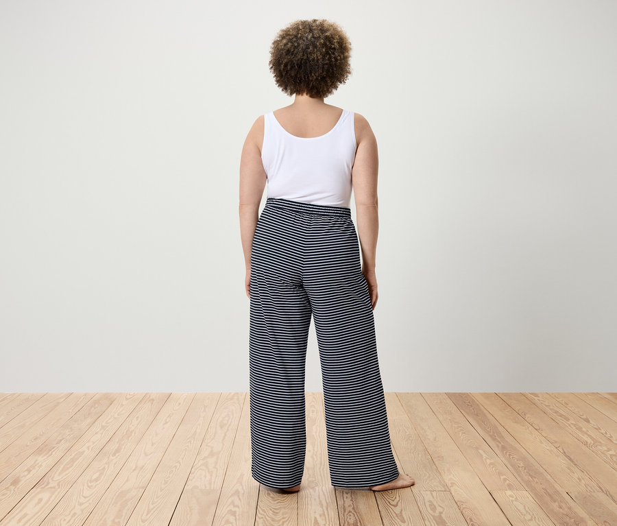 Rückansicht einer Frau mit lockigem Haar in einem weißen Tanktop und gestreiften Hosen.