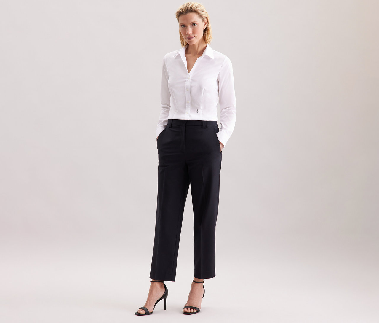 Frau steht und trägt eine weiße Seidensticker Bluse Slim Fit non iron, dazu eine schwarze Hose und schwarze High Heels.