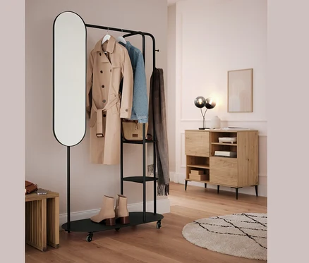 Schwarzer Kleiderständer mit ovalem Spiegel und Ausziehhocker. An der Stange hängen ein beiger Mantel, eine blaue Jacke und ein grauer Schal.