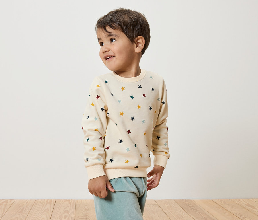 Junge mit sternenverziertem, cremefarbenem Pullover und blauen Samthosen steht auf Holzboden und schaut nach links.