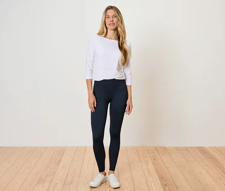 Eine Frau steht auf einem Holzboden und trägt ein weißes Langarmshirt und schwarze Leggings.