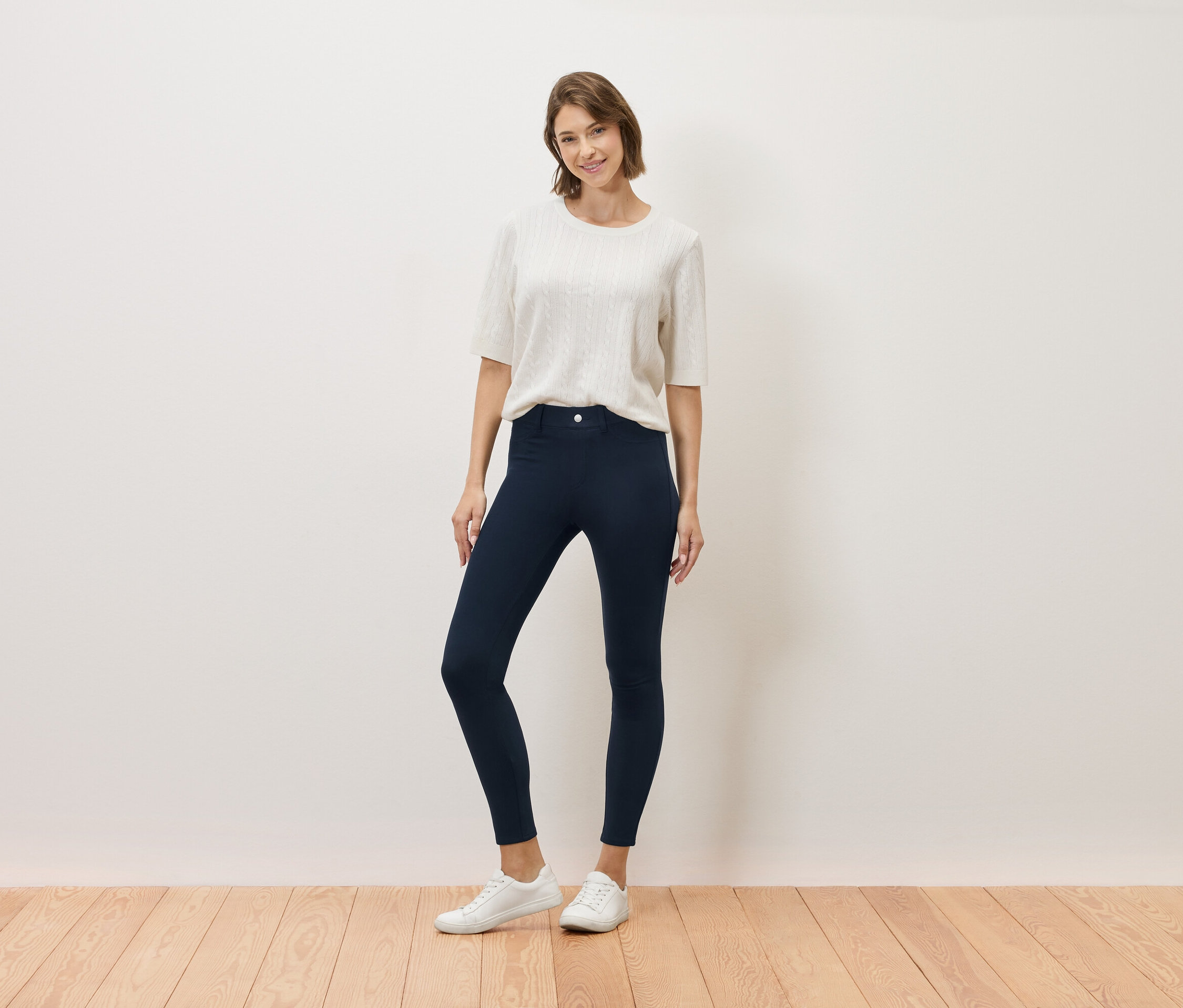 Frau posiert in cremefarbenem Oberteil, marineblauer Hose und weißen Turnschuhen.