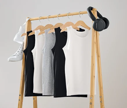 An einer hölzernen Kleiderstange hängen Tanktops in Schwarz, Weiß und Grau. Ein weißer Schuh und schwarze Kopfhörer hängen an der Stange.