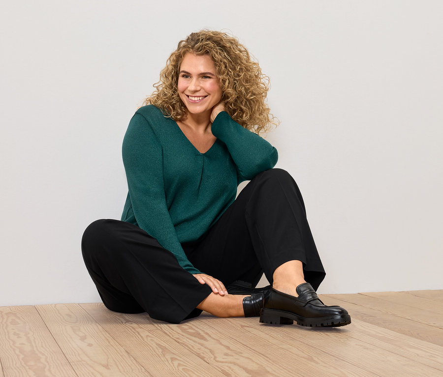 Eine Frau sitzt auf dem Boden und trägt einen grünen Pullover und eine schwarze Hose.