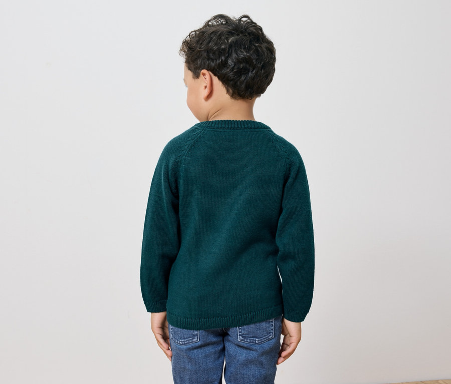 Ein Junge mit dunklen, lockigen Haaren steht mit dem Rücken zur Kamera und trägt einen grünen Pullover und Jeans.