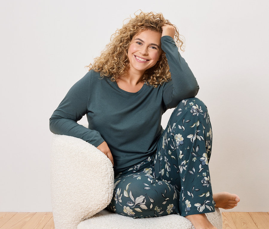 Frau mit lockigem Haar sitzt in einem Sessel und trägt einen dunkelgrünen Schlafanzug mit Blumenmuster.