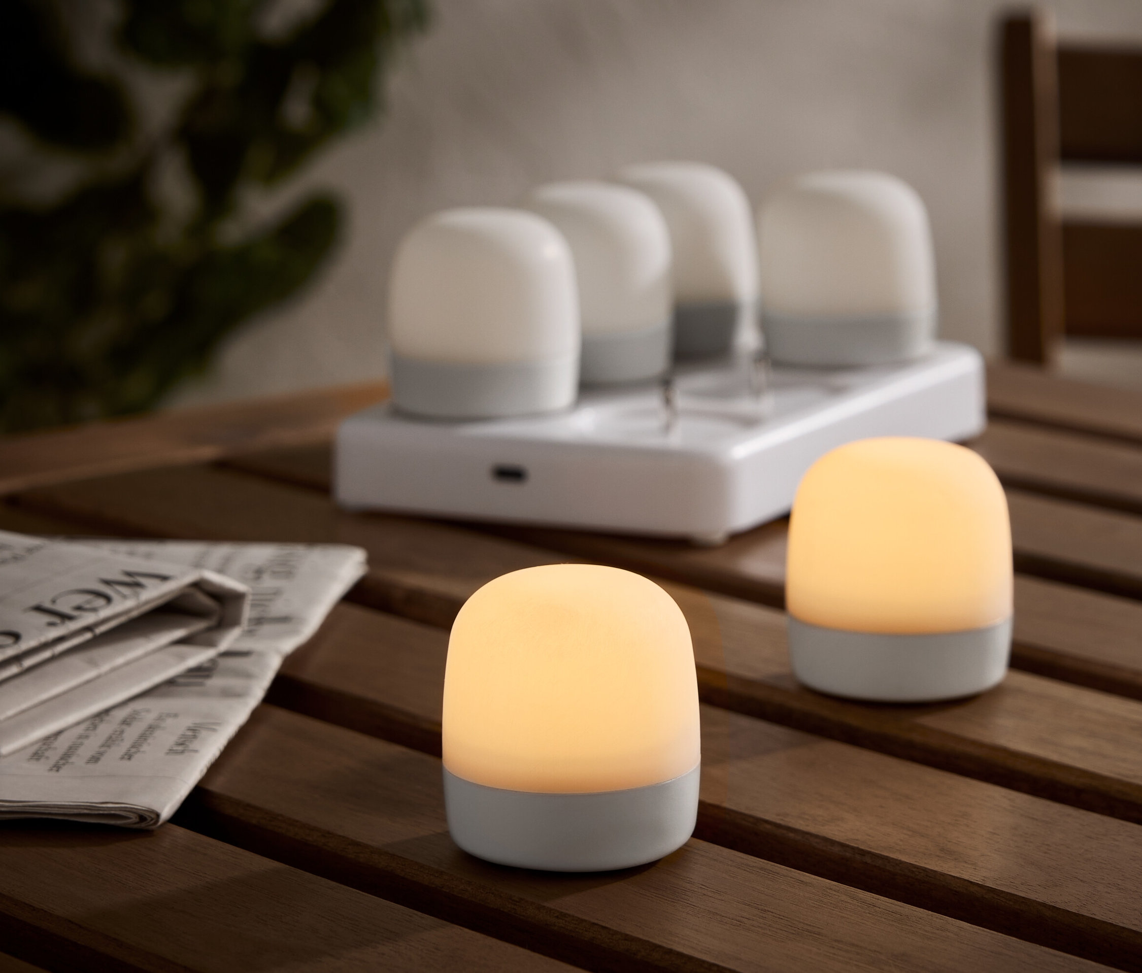 Zwei beleuchtete, kabellose Tischleuchten auf einem Holztisch, mit einer Ladestation und vier weiteren Leuchten im Hintergrund. Eine Zeitung liegt daneben.