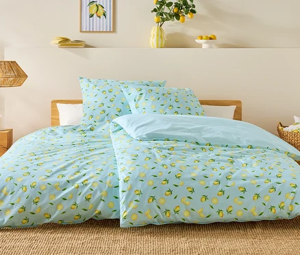 Bett mit hellblauem Bettbezug mit Zitronenmuster, zwei Kissen und einer hellblauen Decke. Auf dem Nachttisch stehen Kerzen.