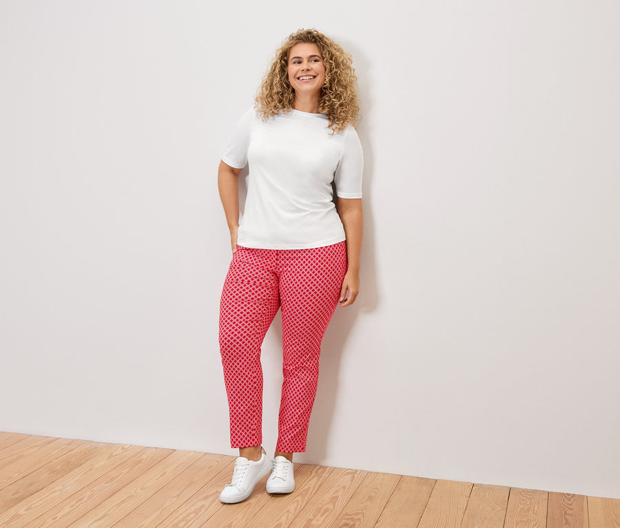 Frau mit lockigem Haar, trägt ein weißes T-Shirt, eine rot gemusterte Hose und weiße Turnschuhe, steht vor einer weißen Wand.