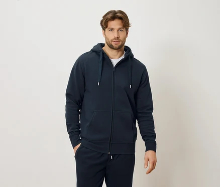Mann mit marineblauem Reißverschluss-Hoodie und marineblauer Jogginghose, stehend vor einem cremefarbenen Hintergrund.