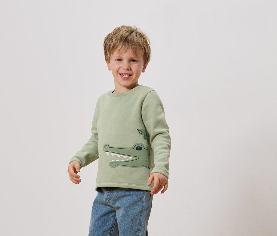 Ein lächelnder Junge trägt ein hellgrünes Sweatshirt mit einem Krokodil darauf und Jeans.
