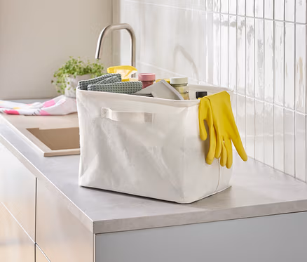 Auf einer Küchentheke steht eine weiße Stoffbox mit verschiedenen Reinigungsutensilien und gelben Gummihandschuhen.
