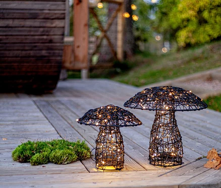 Auf einer Holzterrasse stehen zwei pilzförmige, schwarze Drahtleuchten mit warmweißen Lichtern neben Moos.