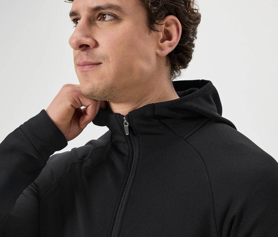 Mann mit lockigem Haar trägt eine schwarze Kapuzenjacke mit Reißverschluss und hält eine Hand an den Kragen.