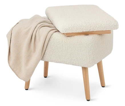 Ein cremefarbener, mit Teddyfell bezogener Hocker mit Holzbeinen und einer beigen Decke liegt darüber.
