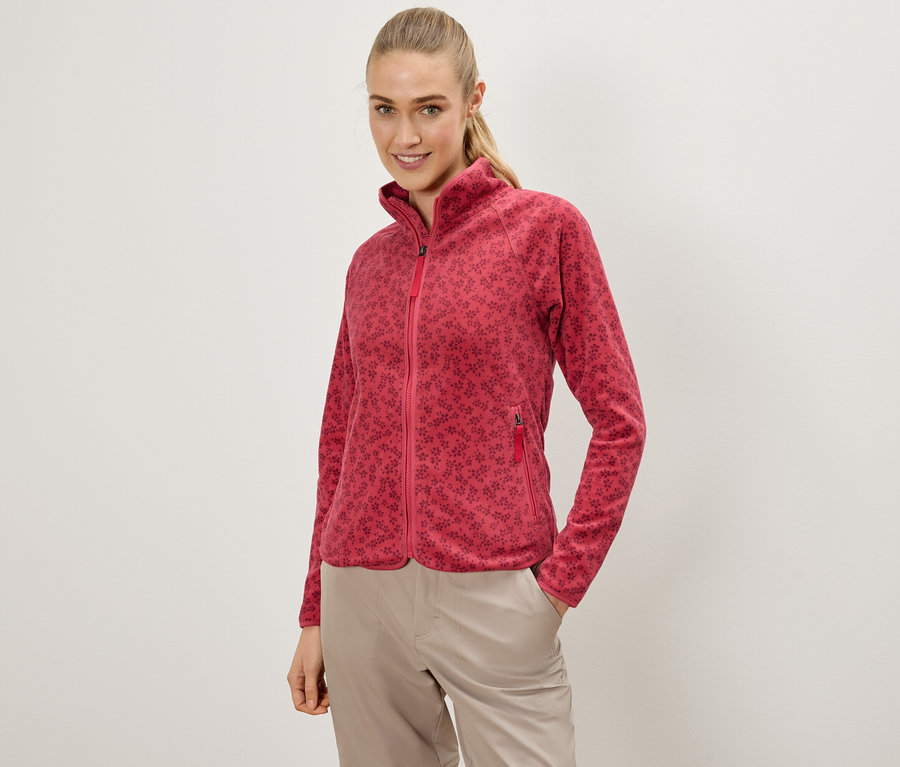 Frau mit einer roten Fleecejacke mit Blumenmuster und beigen Hosen, die mit einer Hand in der Tasche posiert.