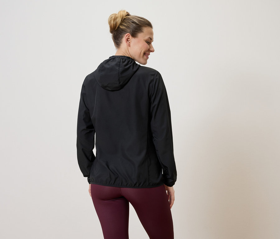 Frau von hinten in schwarzer Kapuzenjacke und weinroter Leggings vor hellem Hintergrund.