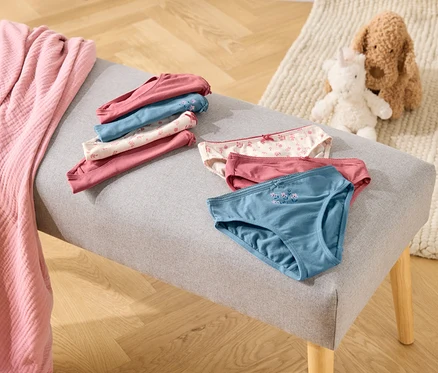 Auf einer grauen Bank liegen gefaltete und ungefaltete Unterhosen in Rosa-, Blau- und Weißtönen, daneben eine rosa Decke und Stofftiere.