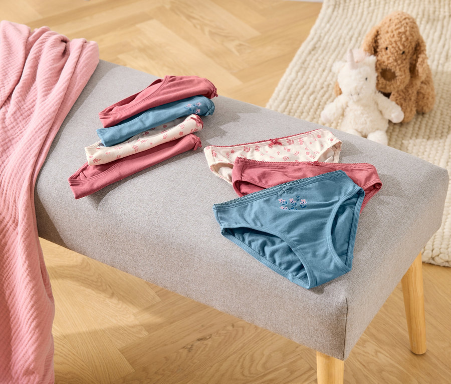 Auf einer grauen Bank liegen gefaltete und ungefaltete Unterhosen in Rosa-, Blau- und Weißtönen, daneben eine rosa Decke und Stofftiere.