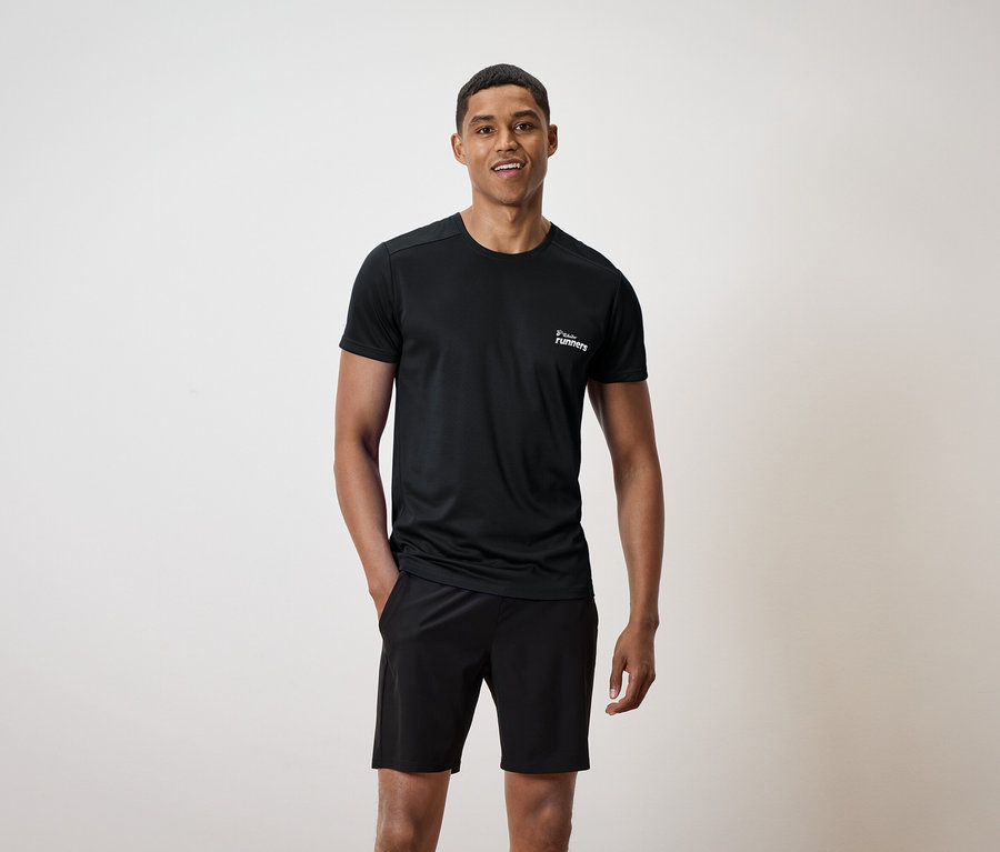 Mann mit schwarzem T-Shirt mit Aufdruck und schwarzen Shorts, eine Hand in der Hosentasche.