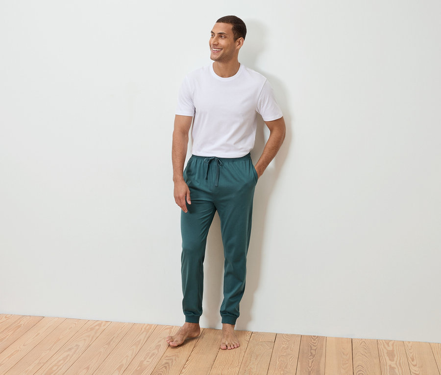 Ein Mann steht barfuß mit der Hand in der Tasche vor einer weißen Wand. Er trägt ein weißes T-Shirt und eine petrolfarbene Jogginghose.
