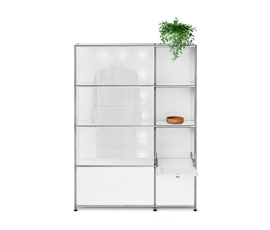 Weißes modulares Regal mit Chromrahmen. Links drei perforierte weiße Paneele und ein geschlossenes Fach unten. Rechts offene Fächer; oben eine grüne Topfpflanze, auf dem dritten Regal eine Holzschale, unten eine teilweise geöffnete Schublade.