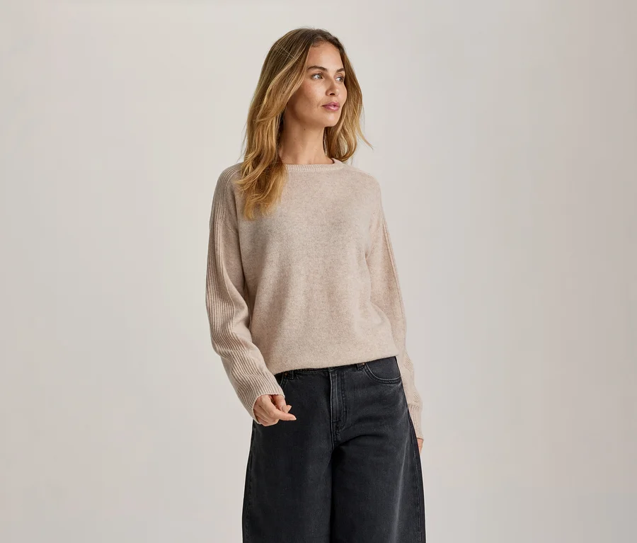 Eine Frau mit langen, welligen blonden Haaren steht vor einem hellen Hintergrund und blickt nach rechts. Sie trägt einen hellbeigen Rundhalspullover mit gerippten Ärmeln und eine dunkelgraue, weit geschnittene Jeans.