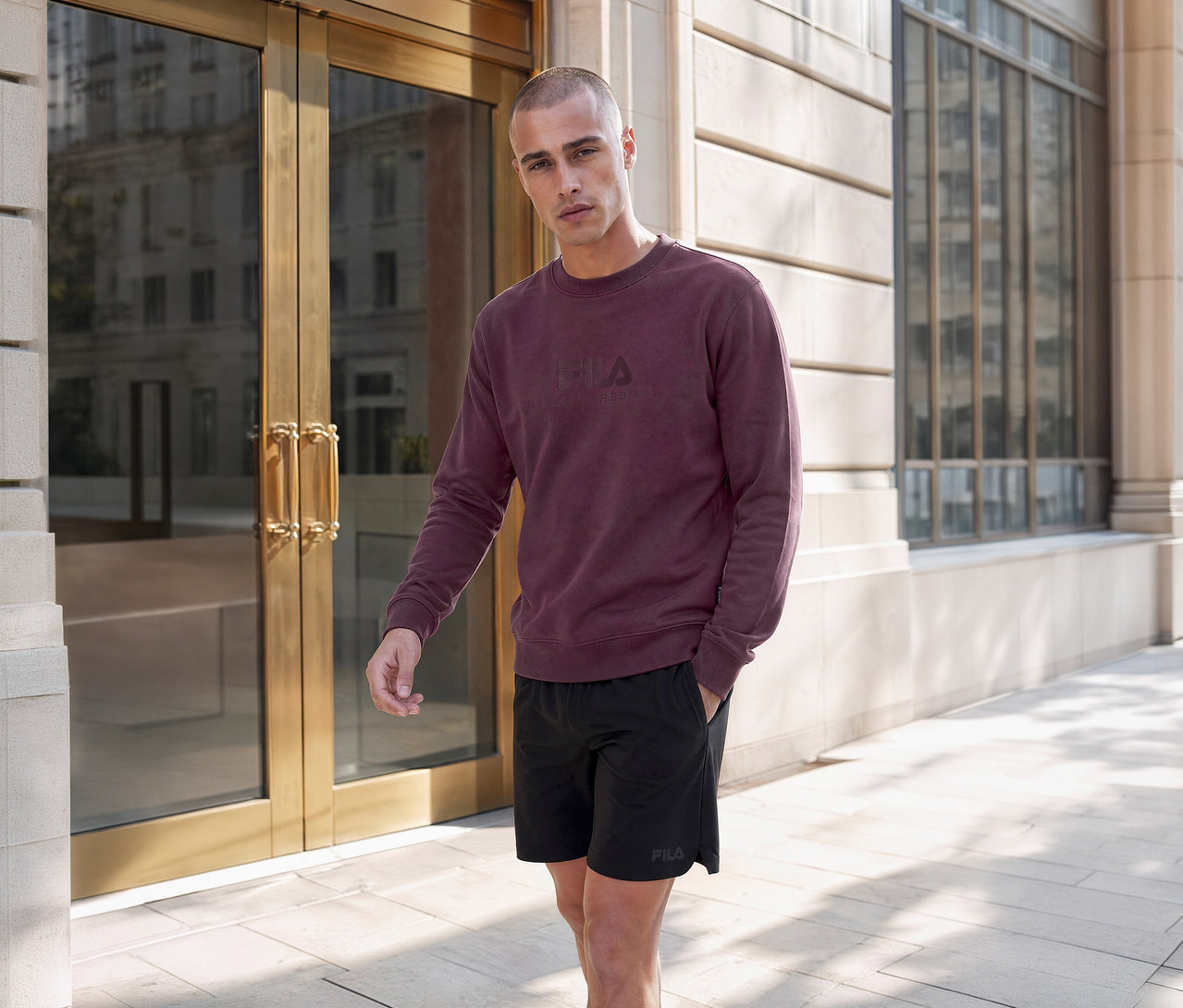 Mann in einem bordeauxroten Fila-Sweatshirt und schwarzen Fila-Shorts steht vor einem modernen Gebäude mit Glastüren.