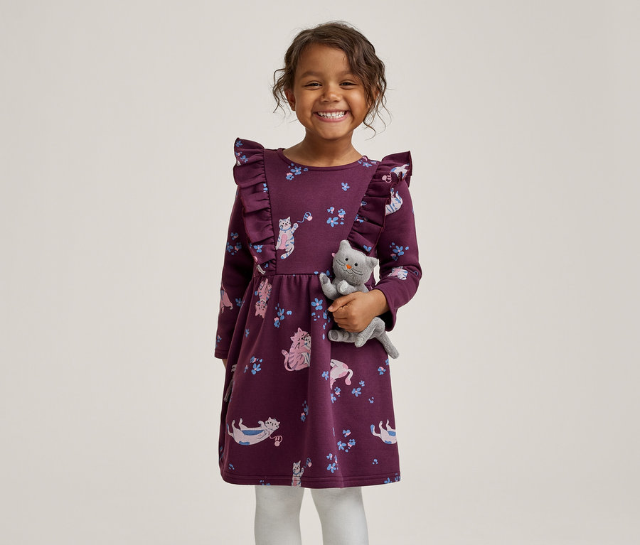 Ein lächelndes kleines Mädchen in einem lila Kleid mit Rüschen und Katzenmuster, das ein graues Plüschkätzchen hält.