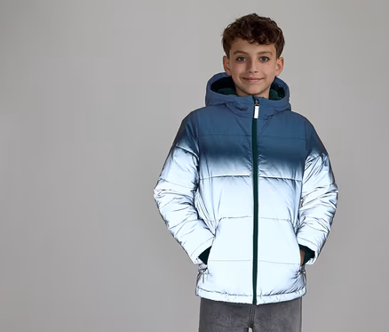 Ein lächelnder Junge mit lockigem Haar trägt eine Kapuzen-Steppjacke. Die Jacke hat einen Farbverlauf von Dunkelblau oben zu reflektierendem Silber unten und einen grünen Reißverschluss. Seine Hände stecken in den Jackentaschen.