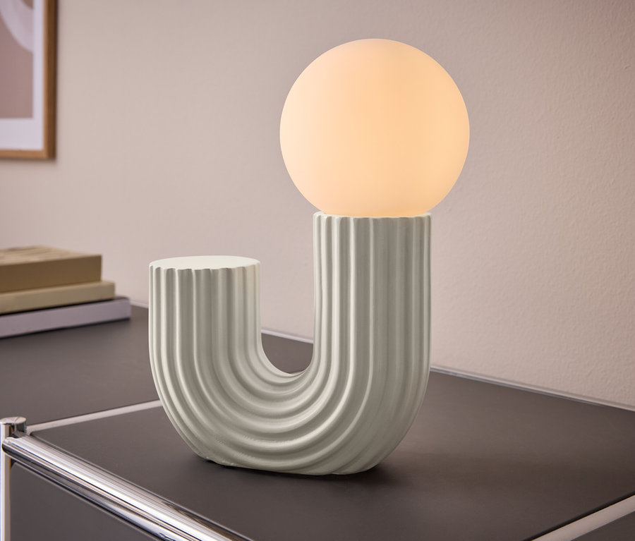 U-förmige, gerippte Tischlampe mit einer leuchtenden Kugel auf einem dunklen Schrank. Im Hintergrund sind Bücher und ein Bild an der Wand.