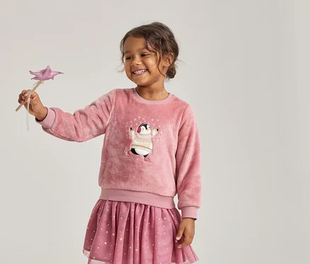 Lächelndes Mädchen in einem flauschigen rosa Pullover mit Pinguin-Motiv und einem rosa Rock mit Sternen, das einen rosa Sternenzauberstab hält.
