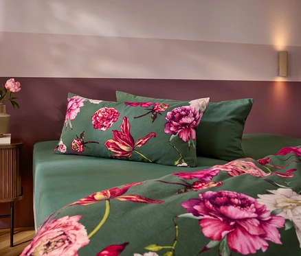 Ein Bett mit dunkelgrüner Bettwäsche, die mit großen rosa und roten Blumen gemustert ist, und einem einfarbigen grünen Kissen. Links steht ein Nachttisch mit einer Vase voller rosa Pfingstrosen. Die Wand ist zweifarbig in Rosa und Dunkelrot gestrichen.
