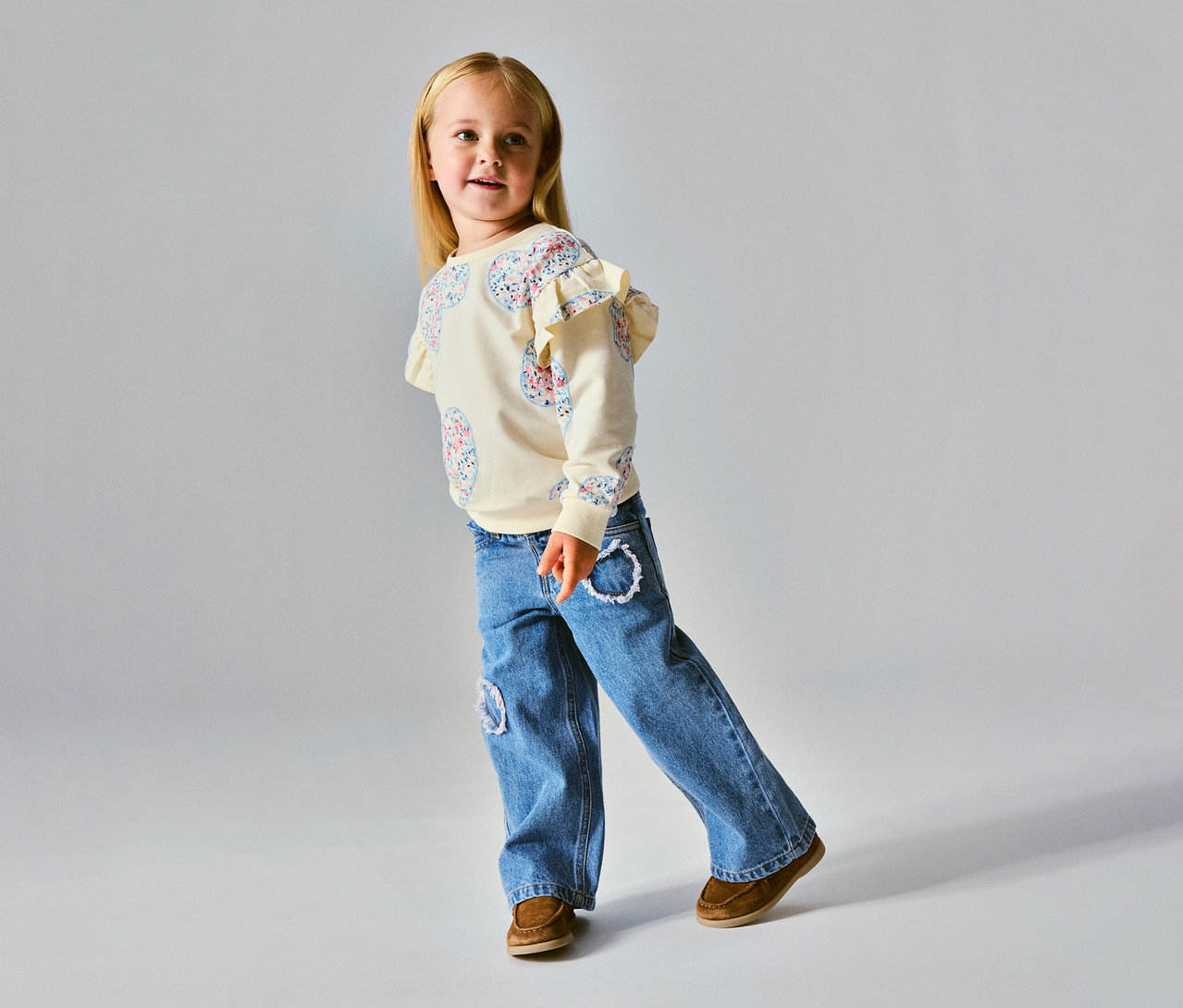 Ein junges Mädchen mit blonden Haaren lächelt, trägt ein cremefarbenes Langarmshirt mit Rüschen an den Schultern und Blumenmuster, blaue Jeans mit weißen Stickereien und braune Mokassins.
