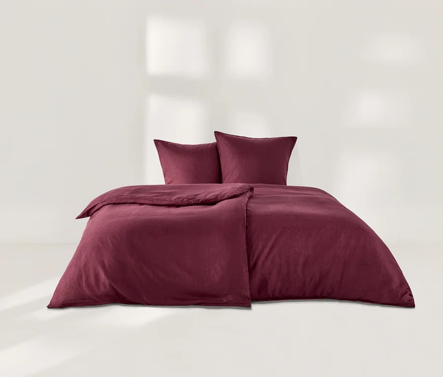 Ein Bett mit beerenfarbener Leinen-Bettwäsche, bestehend aus einem Bettbezug und zwei Kissenbezügen, vor einem weissen Hintergrund.