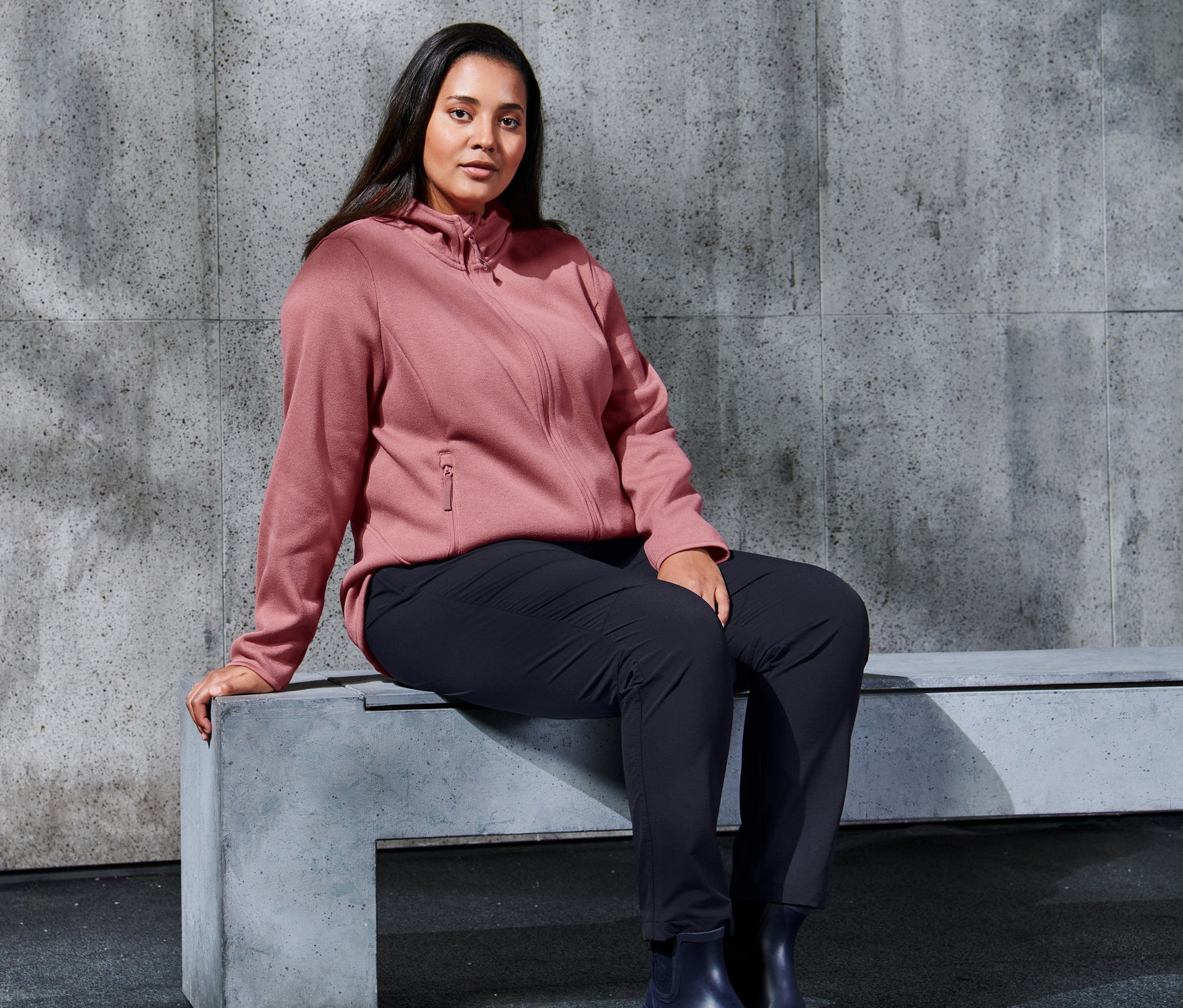 Eine Frau sitzt auf einer Bank und trägt eine roséfarbene Strickfleece-Jacke und eine Softshell-Hose.