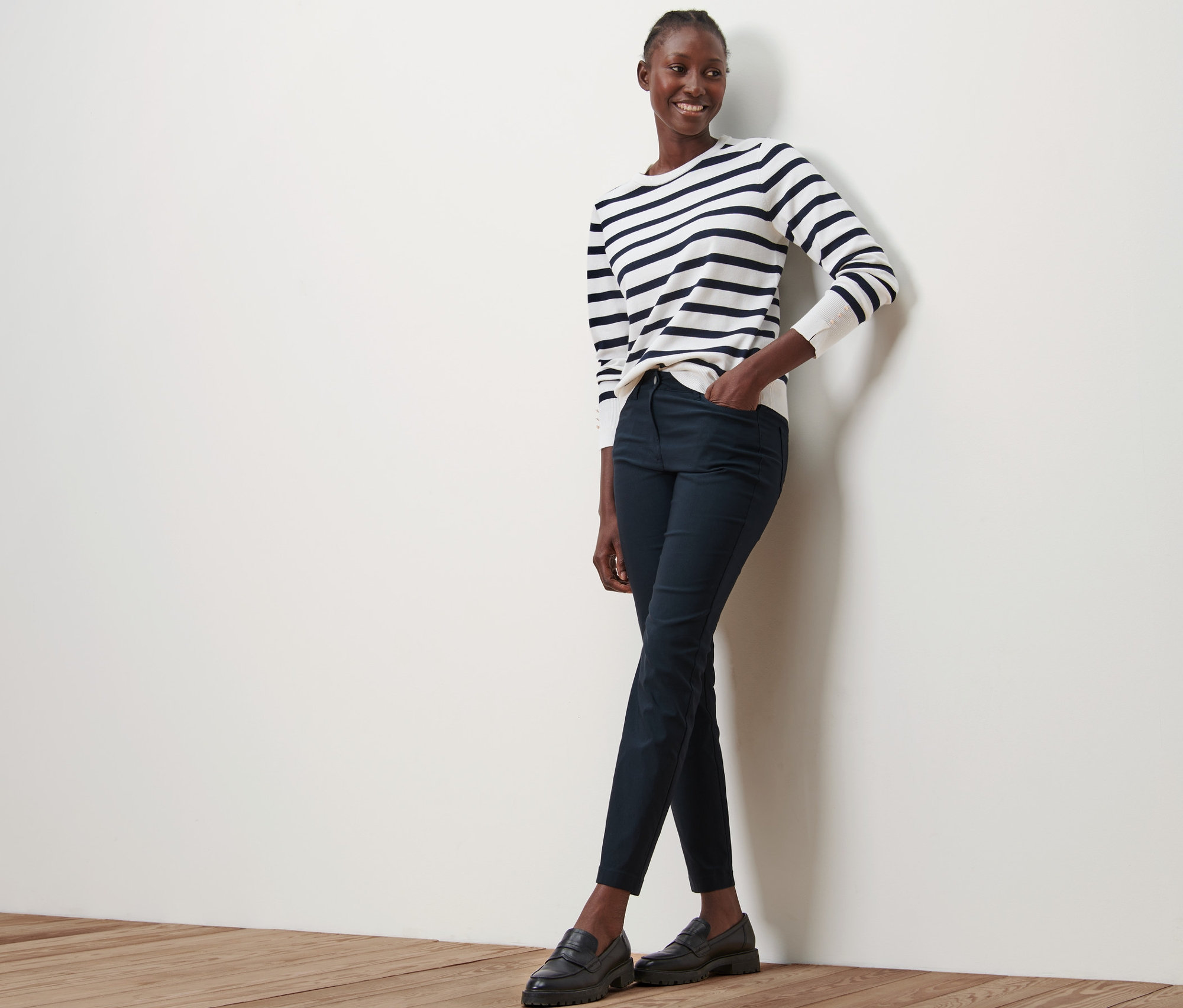 Eine Frau steht an einer Wand und trägt eine dunkelblaue Bengalin-Hose, ein gestreiftes Oberteil und schwarze Schuhe.