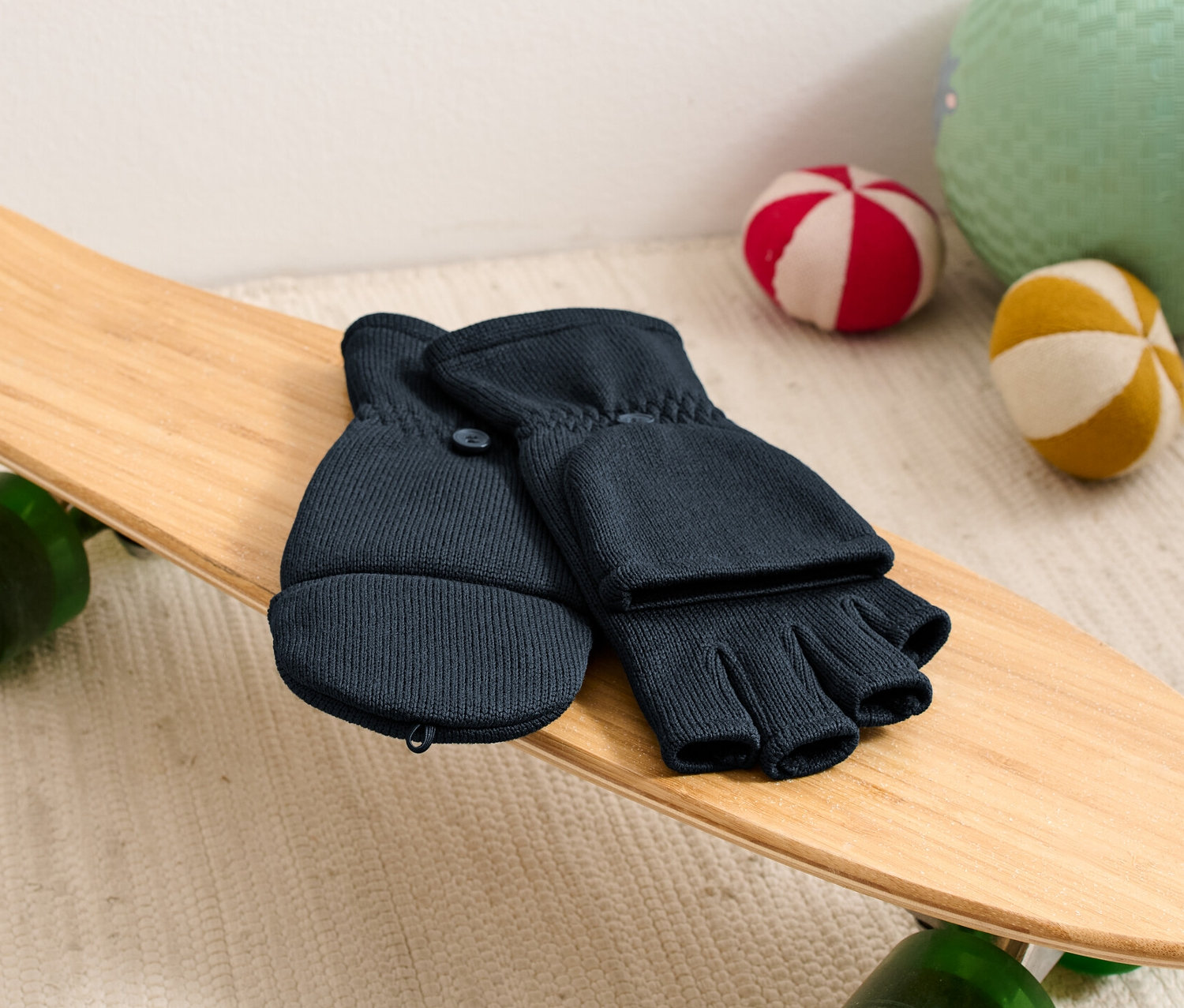 Kinder-Strickfleece-Klappfäustlinge liegen auf einem Skateboard neben Bällen.