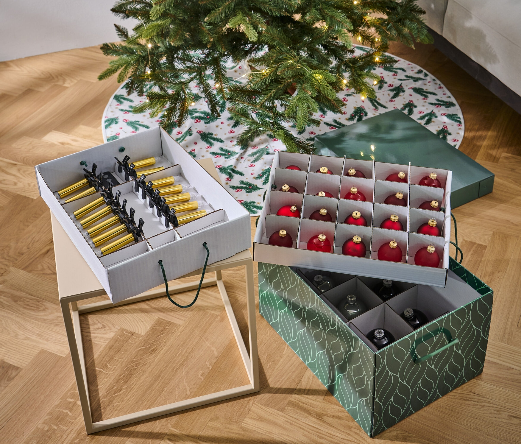 Ein Weihnachtsbaum steht neben zwei Weihnachtsordnungsboxen. Eine Box ist weiß und enthält goldene und schwarze Ornamente, die zweite ist grün und enthält rote und silberne Ornamente.