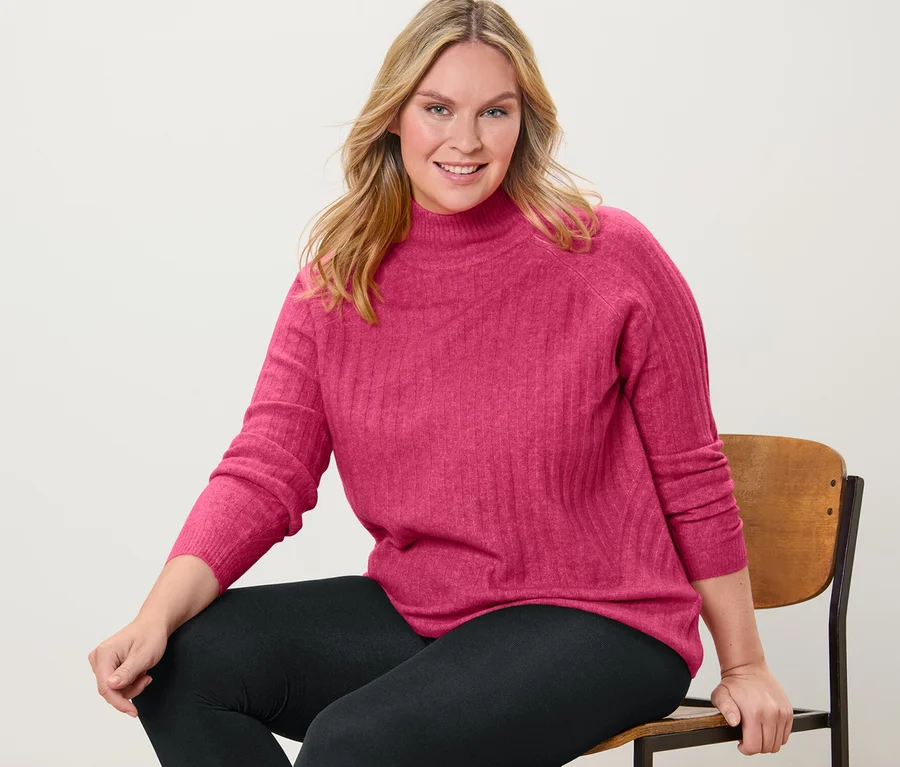 Frau sitzt auf einem Stuhl und trägt einen gerippten Strickpullover mit Stehkragen und Thermoleggings.