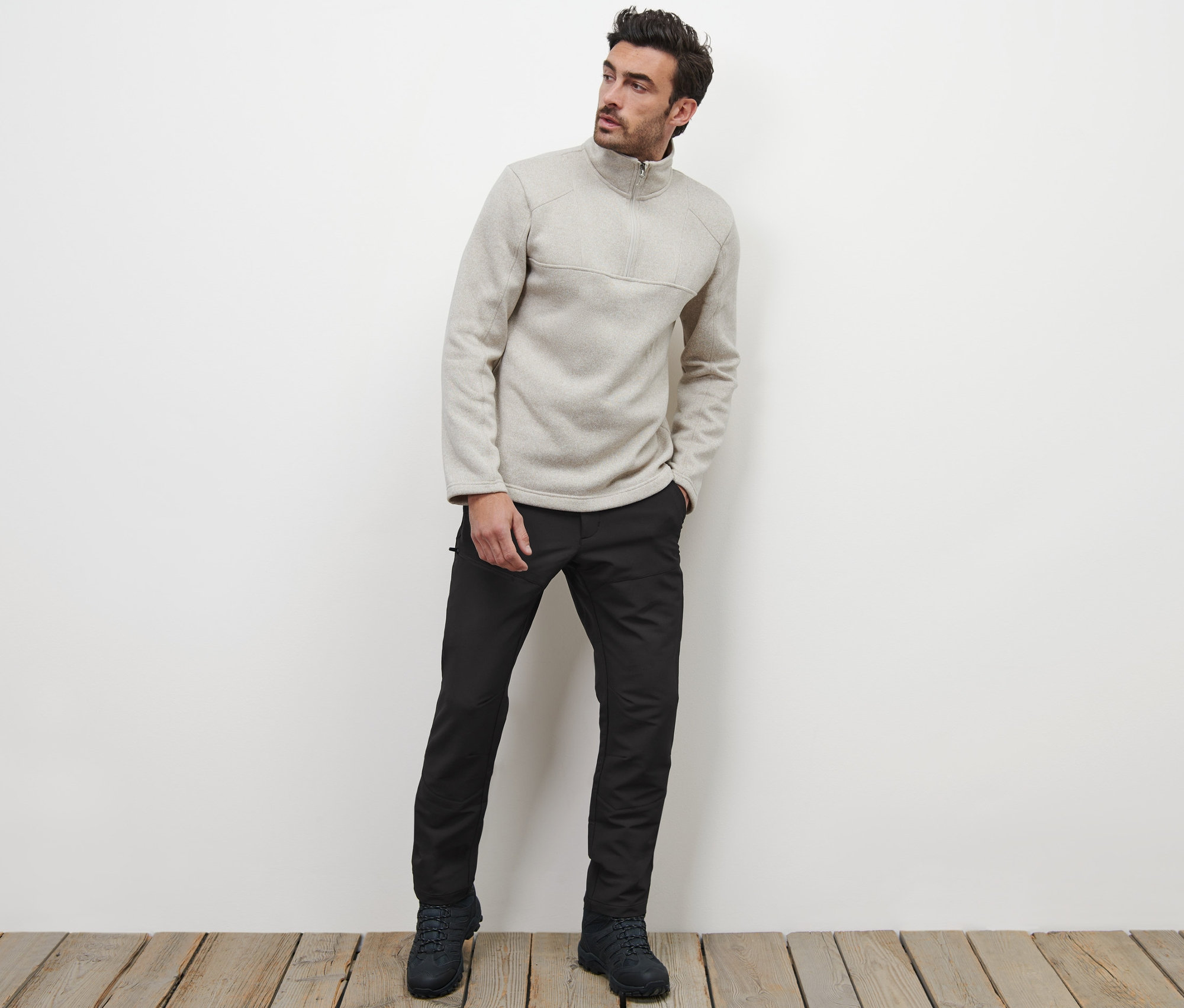 Mann posiert auf einem Holzboden und trägt eine Thermohose und einen Pullover mit Reißverschluss.